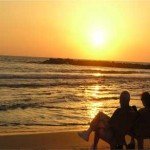 An eldery couple sit on chairs by the beach with a sunset