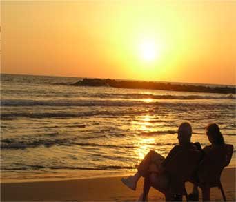 An eldery couple sit on chairs by the beach with a sunset