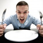 Man holds fork and knife with empty plate and angry look on face