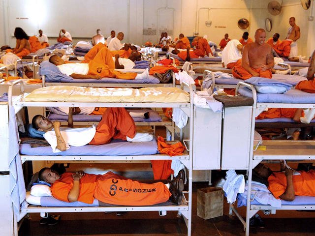 Prison inmates sit or lay in their bunkers wearing orange jumpsuits