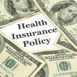 The words Health Insurance Policy surrounded by multiple 100-dollar bills